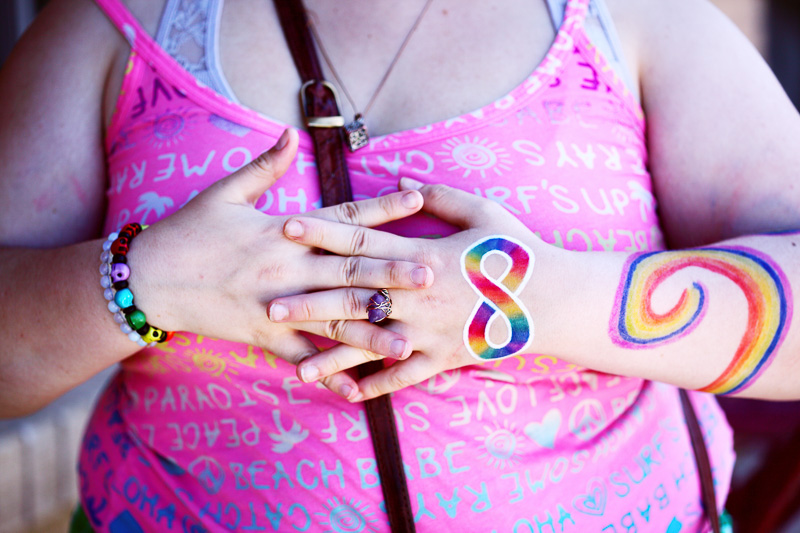 A person wearing a pink top has a rainbow infinity symbol and other rainbow pain on their arms with their hands clasped in front of their chest.